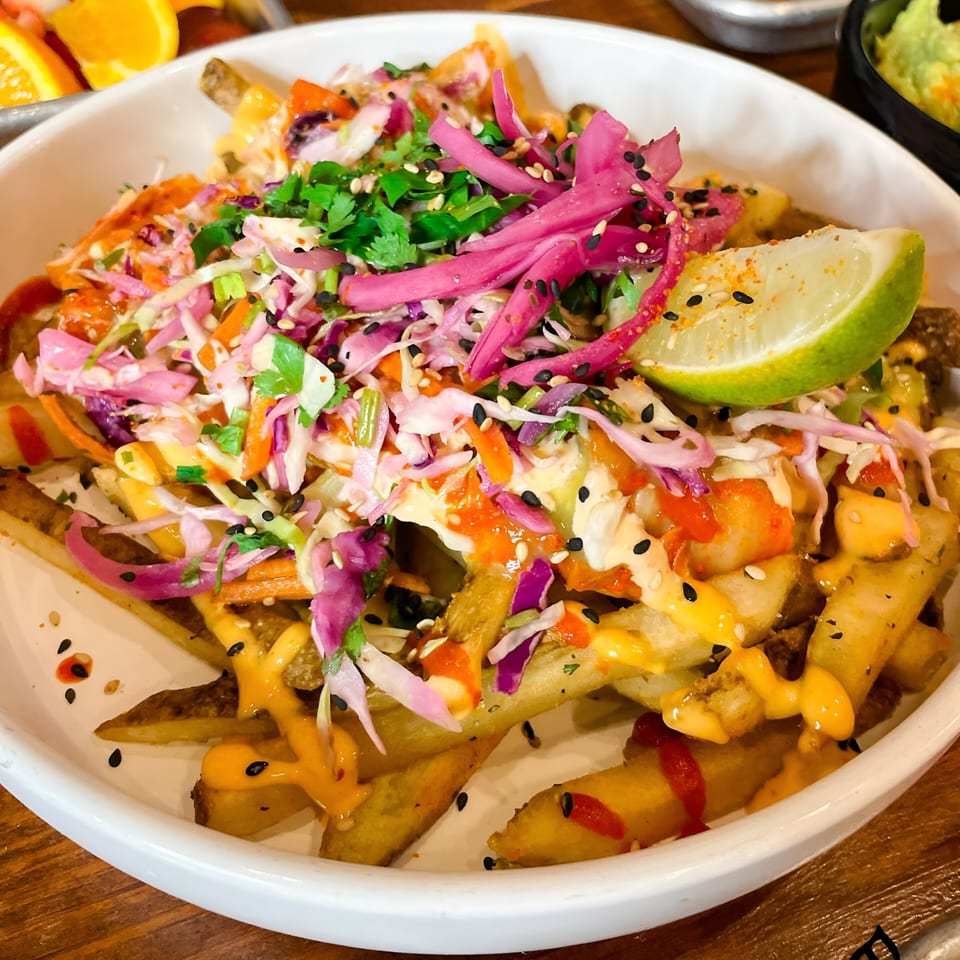 Nuri Fusion Street Kitchen's loaded fries topped with colorful slaw, sauces, sesame seeds, and a lime wedge.