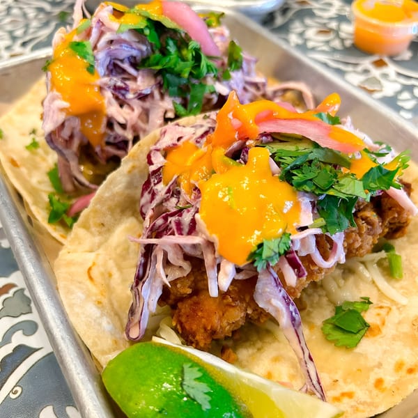 Two Nuri Fusion Street Kitchen tacos with fried chicken, red cabbage slaw, orange sauce, and cilantro.