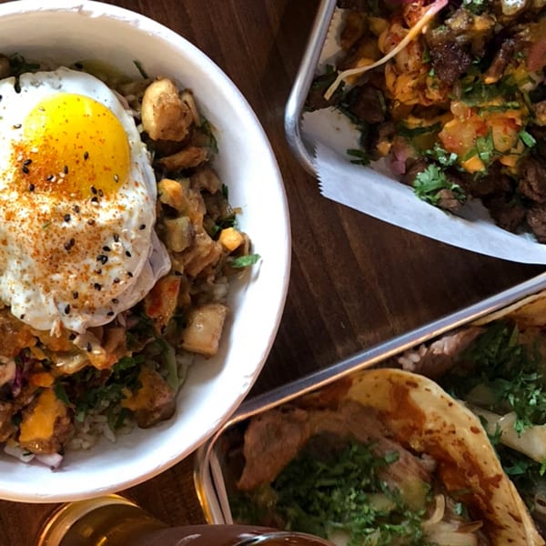 A white bowl with a fried egg over rice and meat, next to two metal trays of tacos.